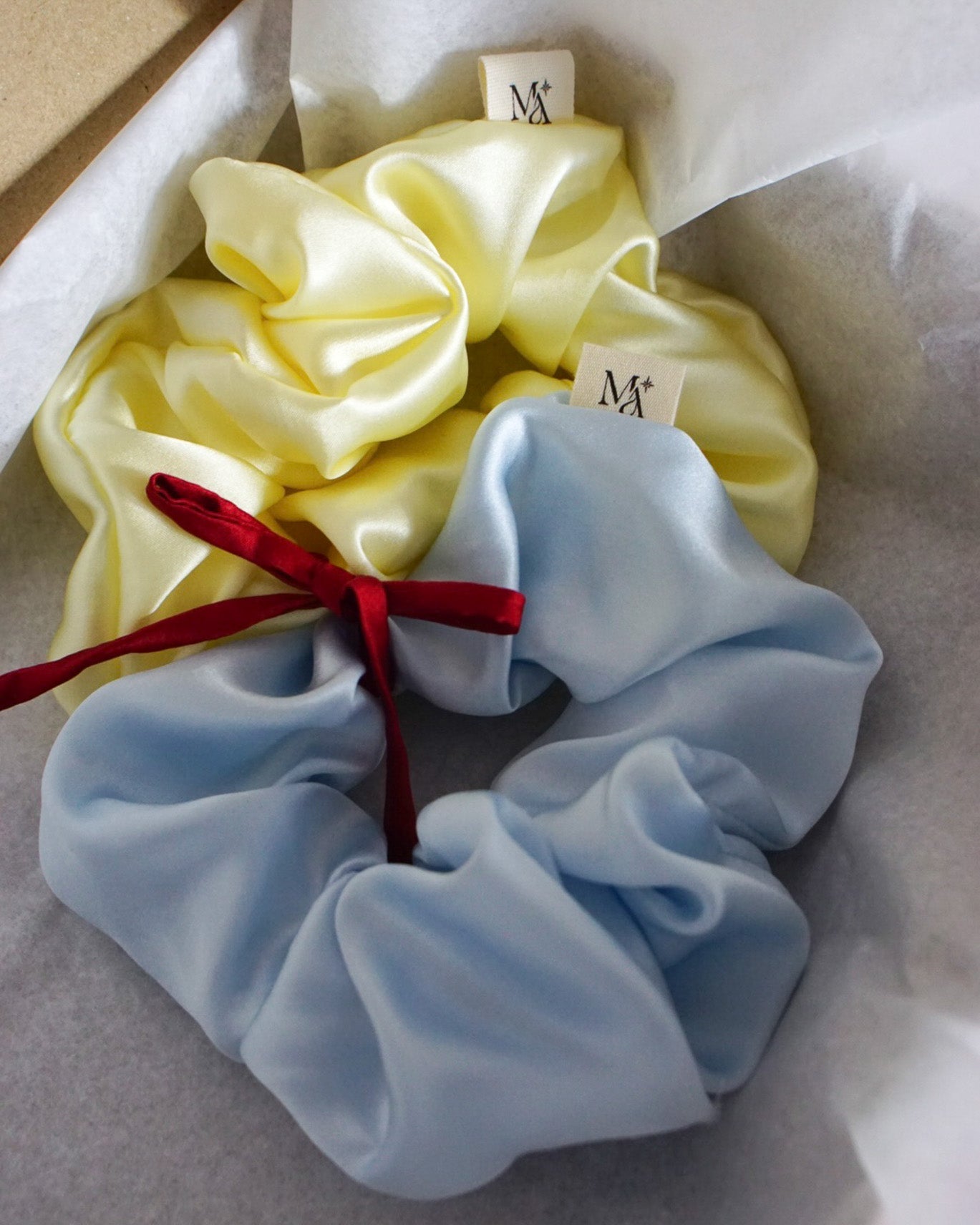 Two silk scrunchies, one yellow and one blue, with a red ribbon on a white background.