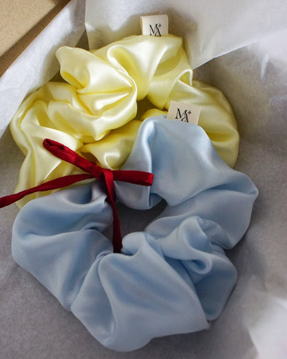 Two silk scrunchies, one yellow and one blue, with a red ribbon on a white background.