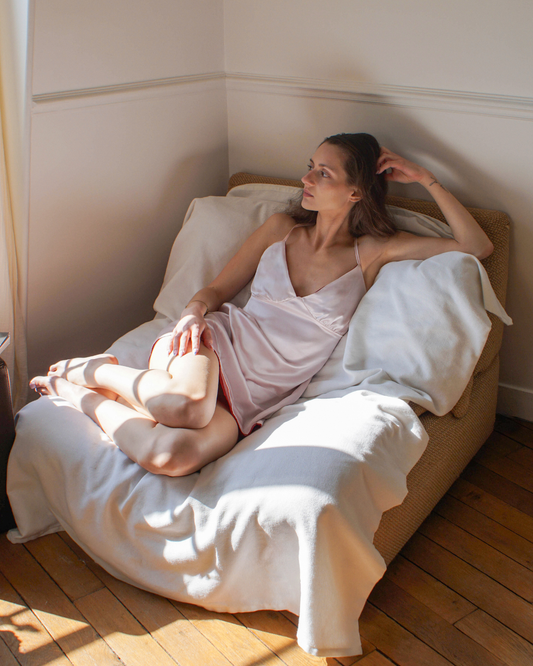 A woman reclining on a bed wearing a pale pink silk dress, surrounded by white bedding and pillows in a sunlit room with wooden floors.